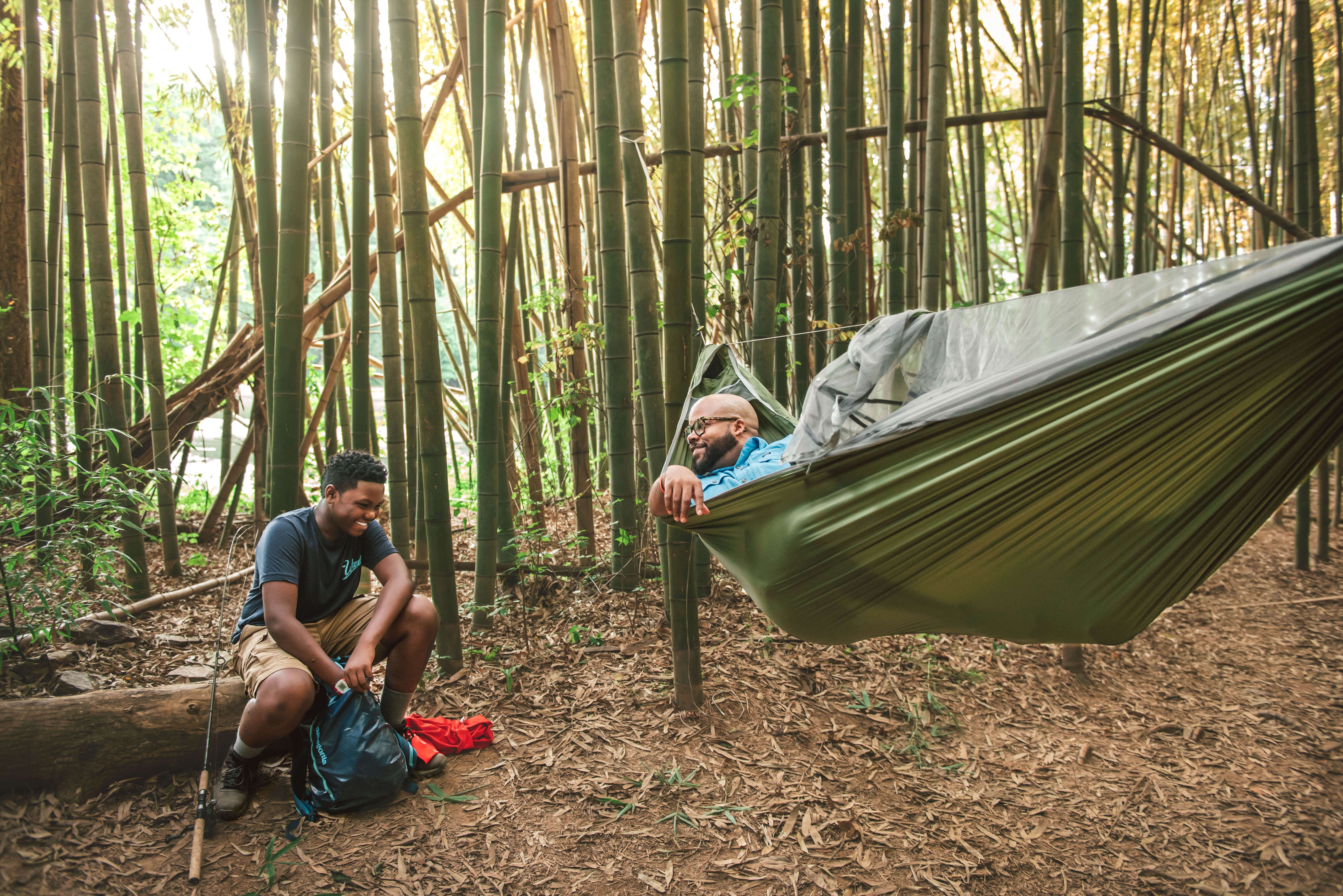 A man in ENO JungleNest hammock looking at another person sitting on the ground. They are in a bamboo forest