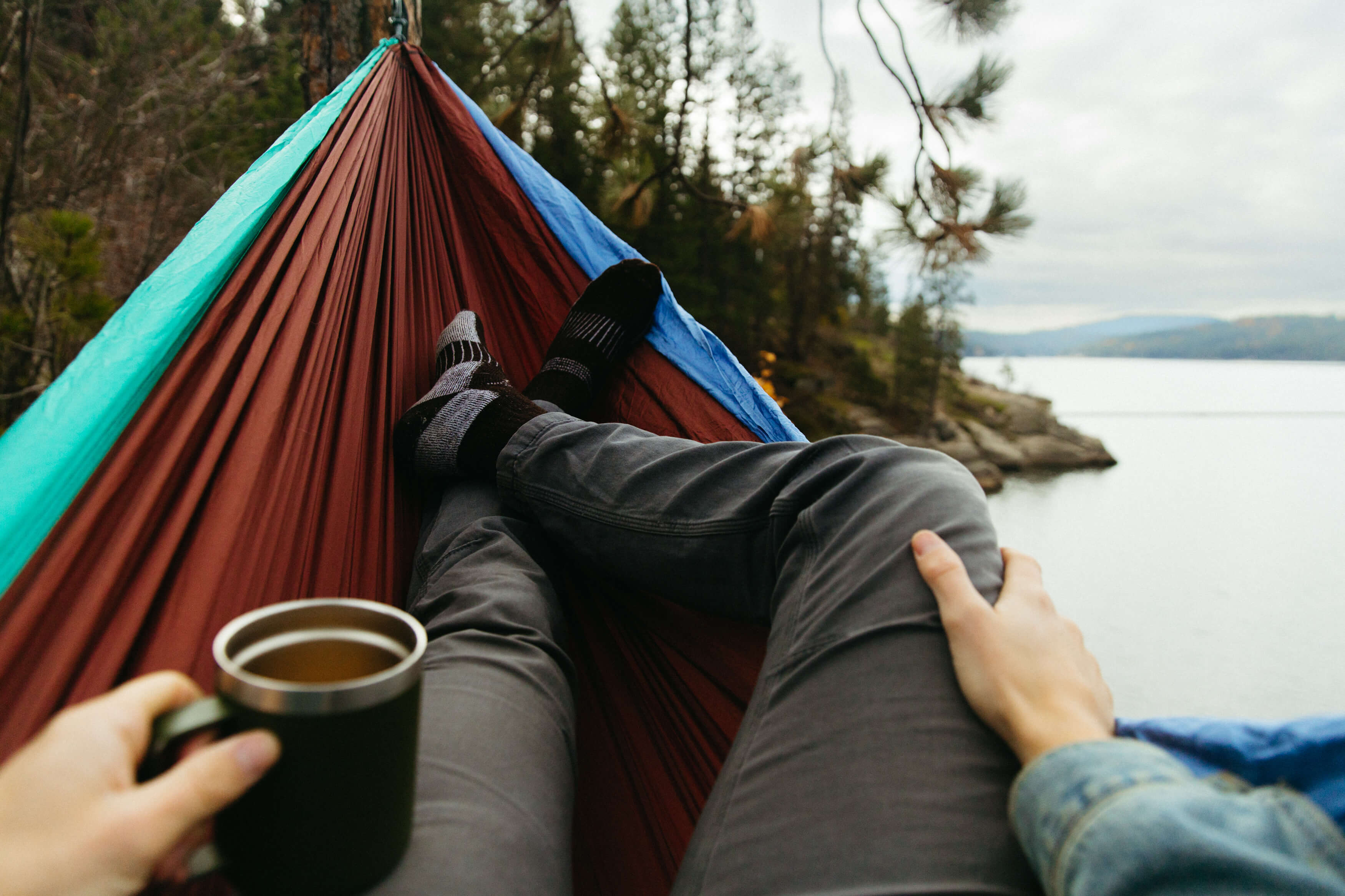 Someones legs in an ENO hammock with coffee looking out at trees and lake