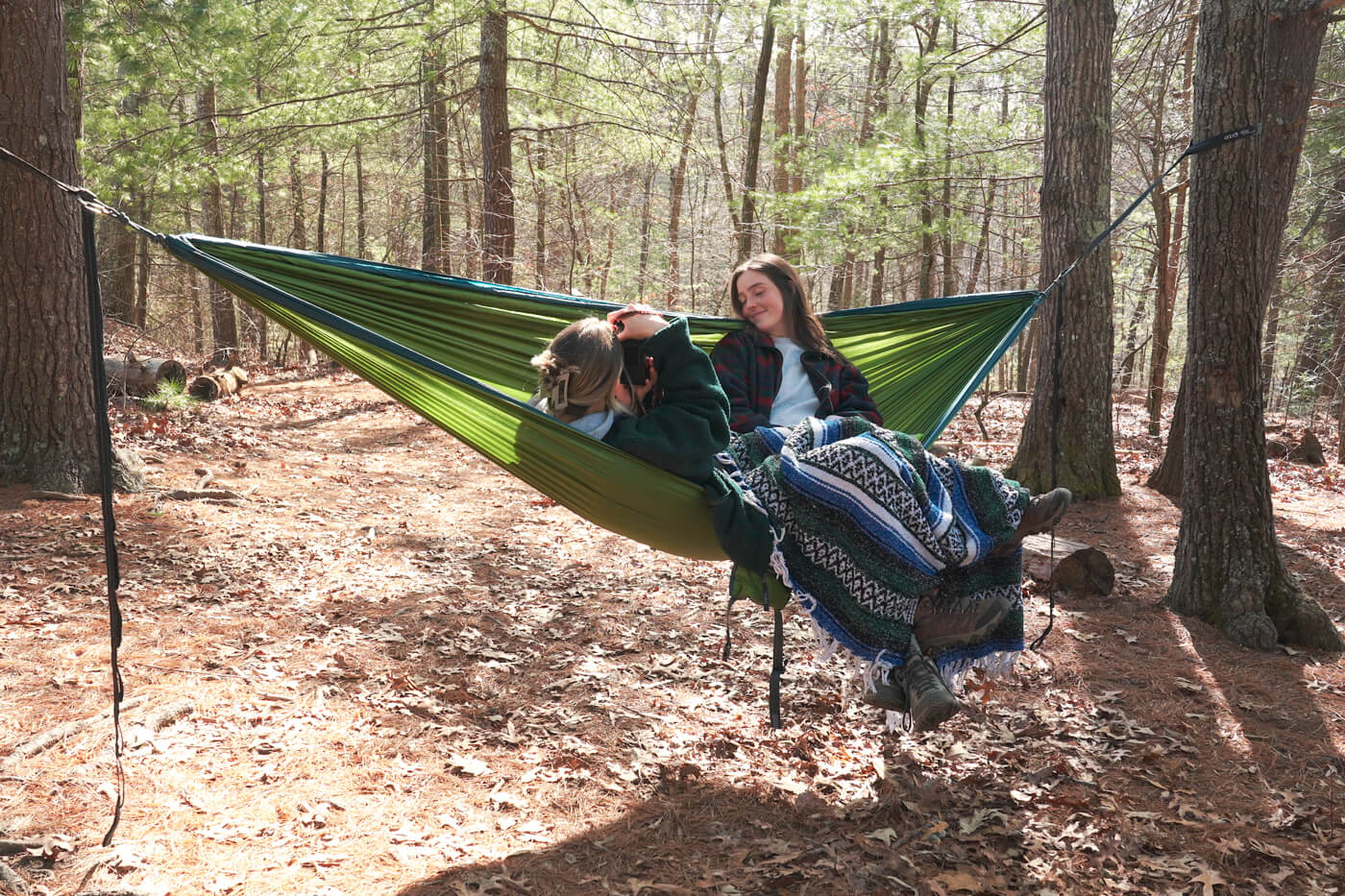A person taking a picture of another person. Both are in an ENO hammock in the woods