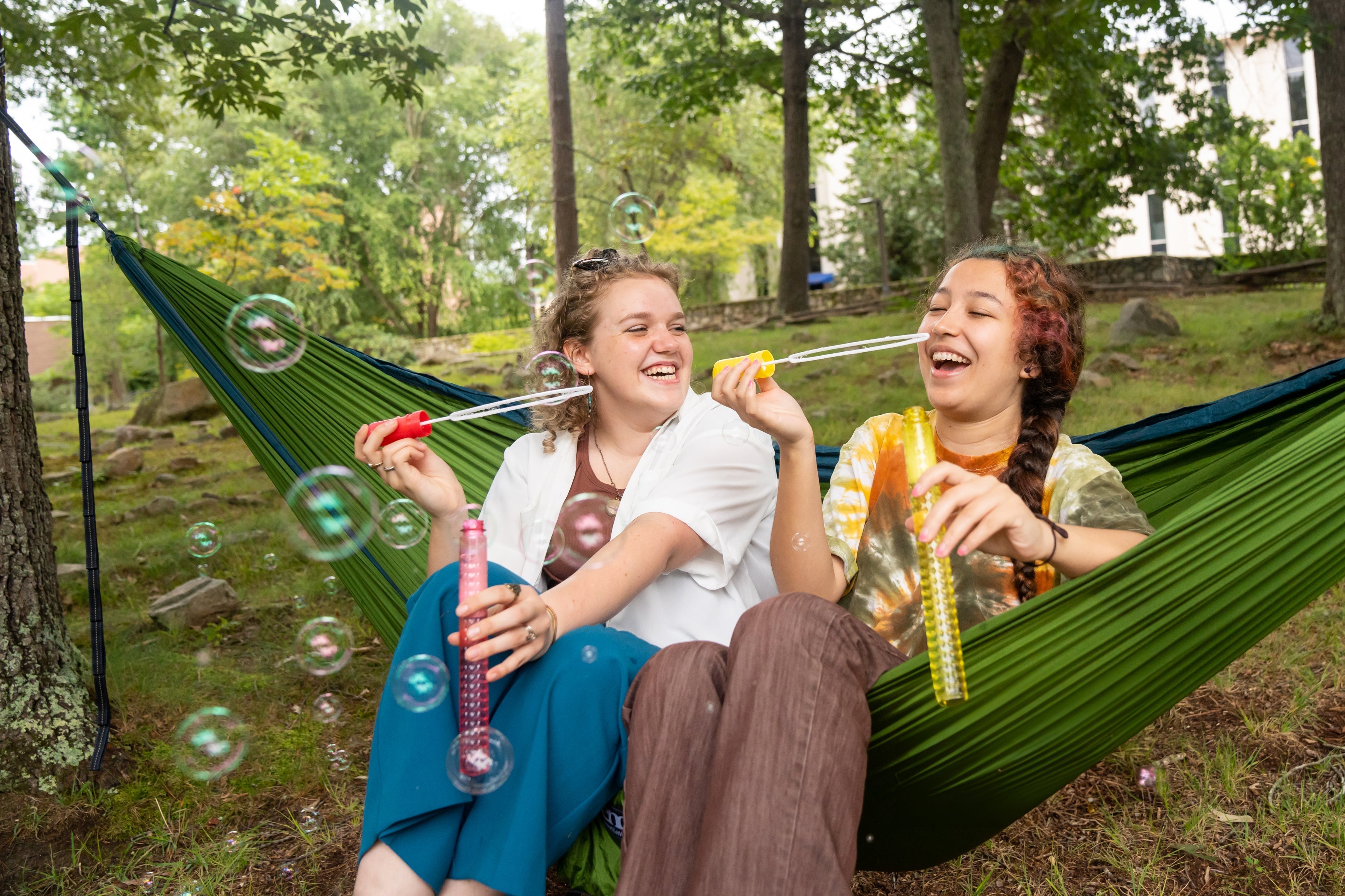 Two girls hammocking in an ENO Hammock together on a college campus while also blowing bubbles