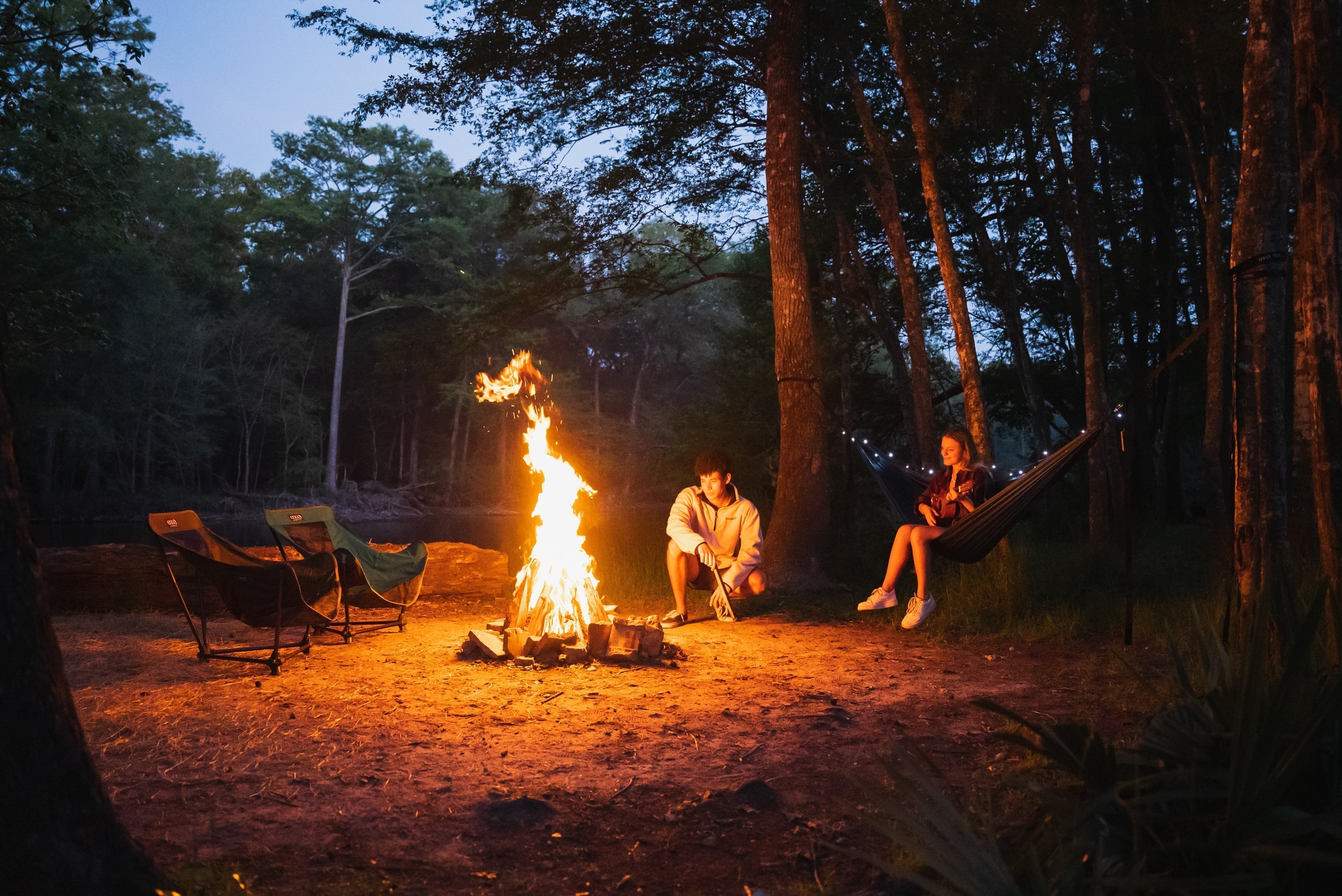 Two people looking at a campfire. One sitting by it and the other is sitting in an ENO Hammock with ENO Twilights strung above