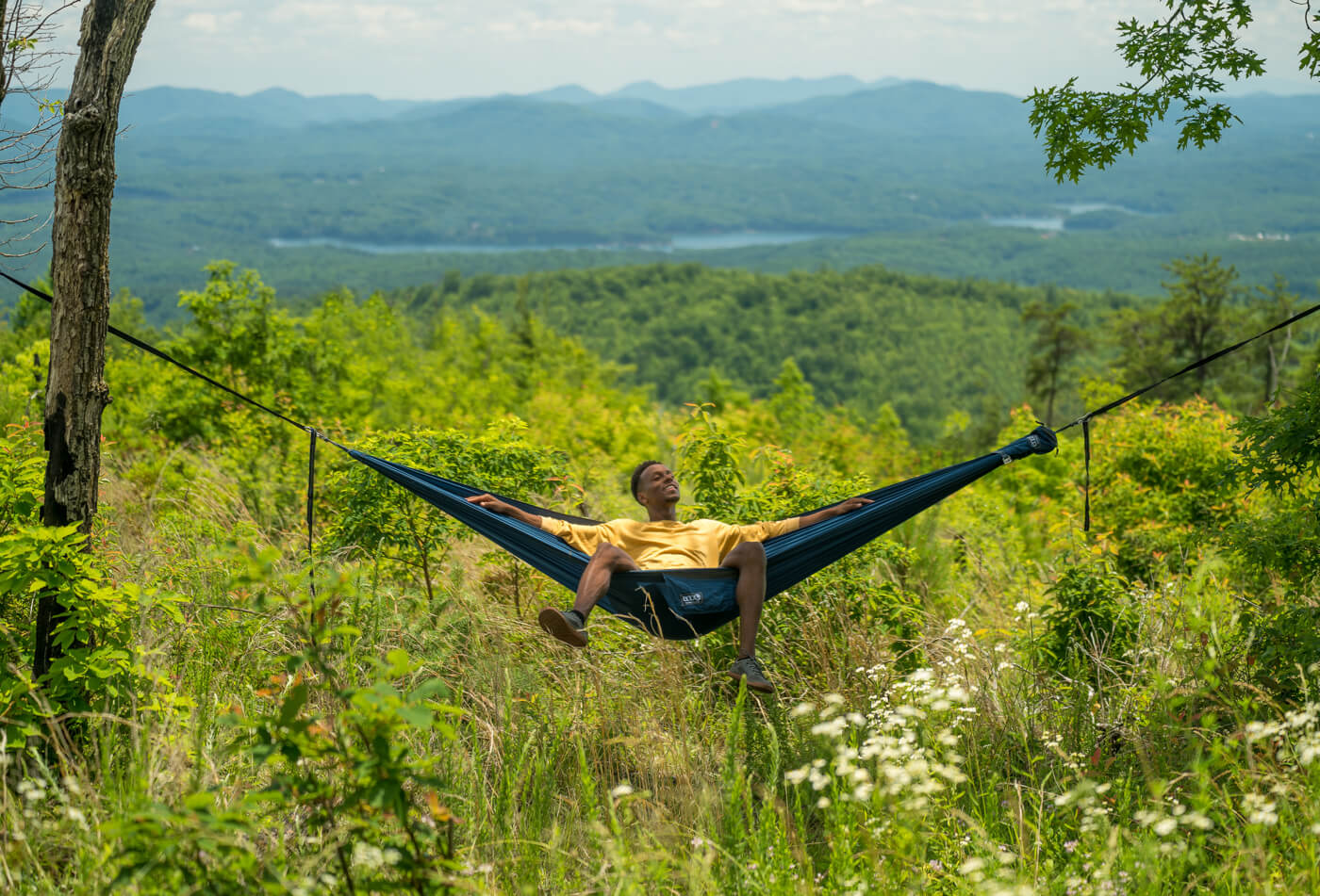 A man sits in an ENO Hammock in a tall field of grass with mountains behind him