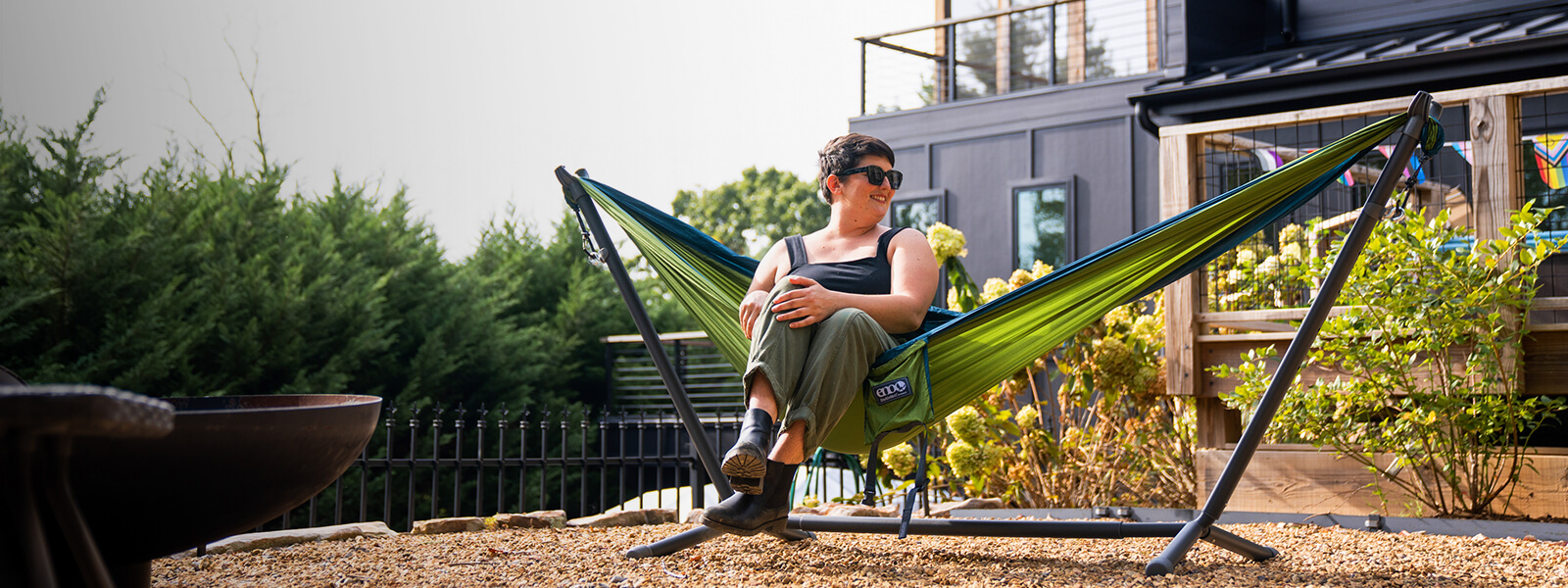 A woman sitting in a ENO portable hammock set up on a portable hammock stand in front of home.