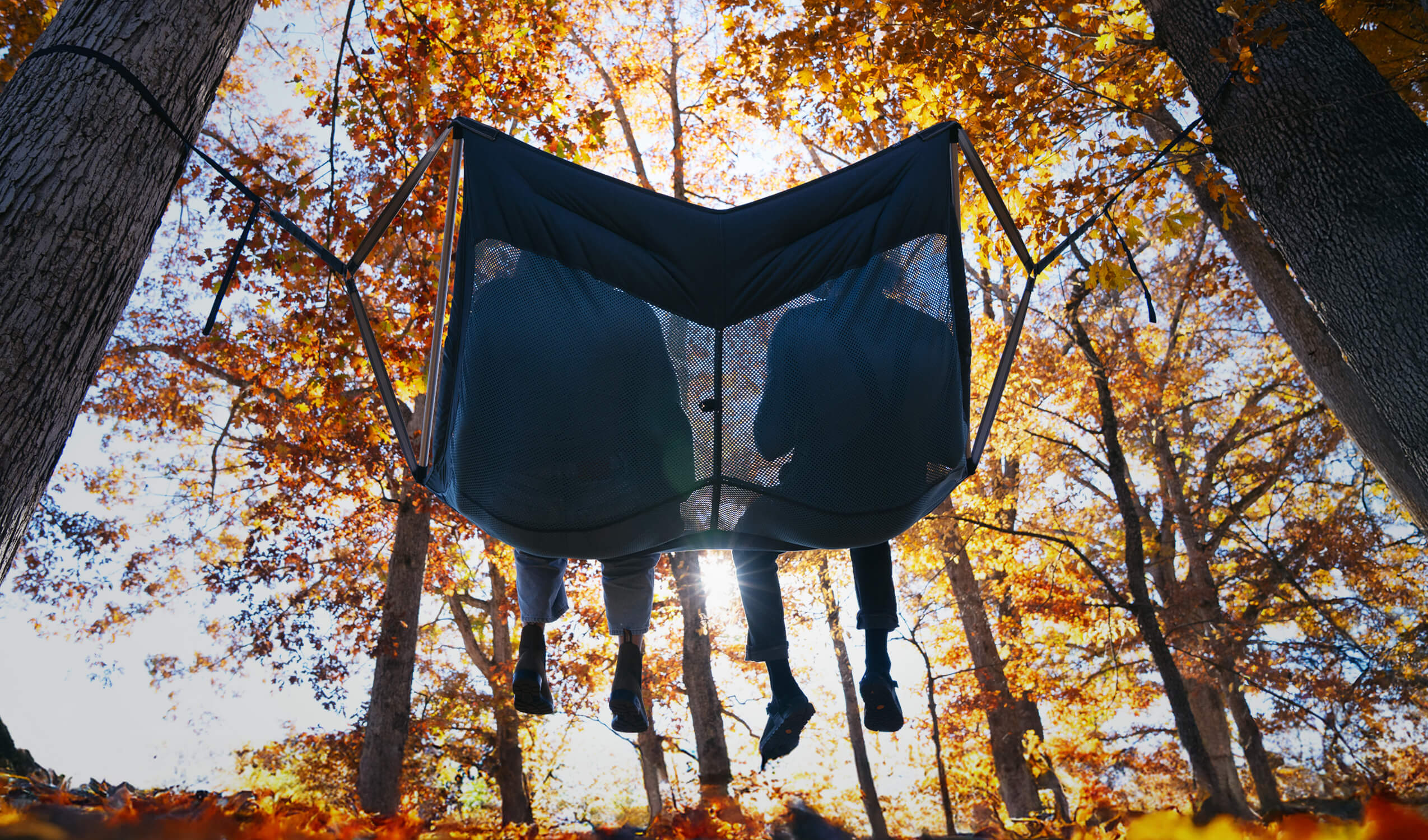 Two people sitting in the DoubleLoft Hammock Chair between trees with autumn foliage
