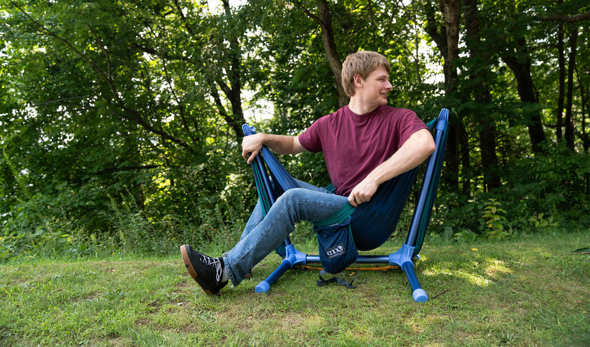 A man sitting outdoors on the ENO Parklite Hammock Chair Stand in new Sky on a grassy lawn with trees in the background.