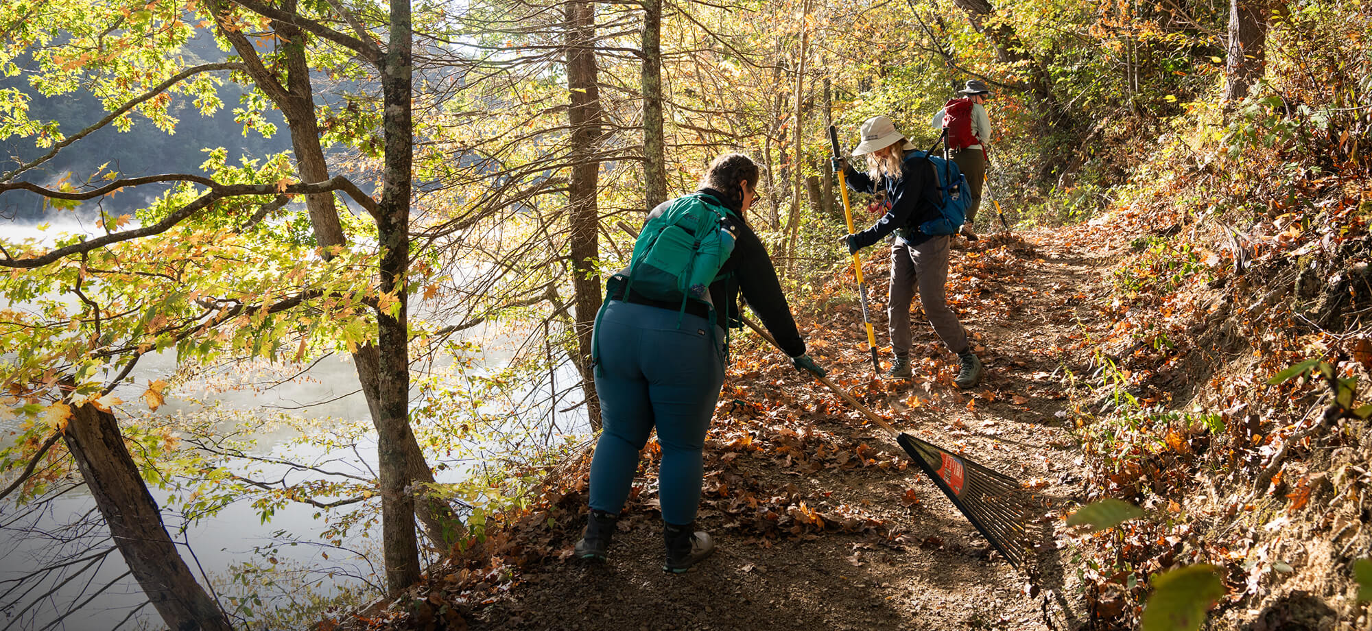 Three hikers on a trail by a lake with trees and autumn foliage.
