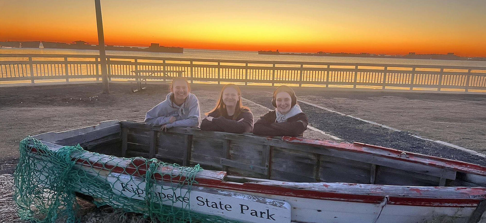 Three people sitting in front of a wooden boat at sunset with a fence and open field in the background.