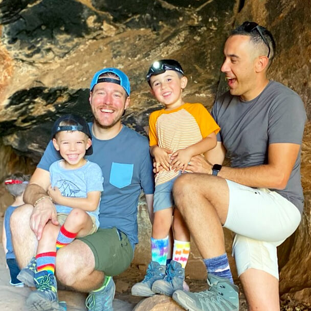 The Fontes Family, comprised of two dads and twin sons,wearing shorts and tshirts laugh while exploring a cave.