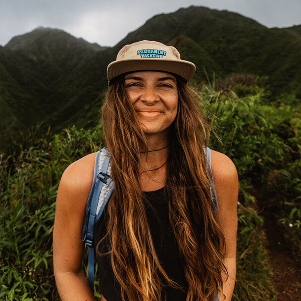 Haleigh Hendrickson, a young woman with long brown hair smiles wearing a basecall cap and backpack while exploring mountains.