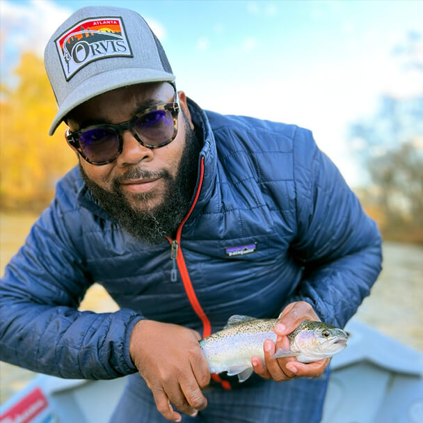 Nick Brooks, DEI advocate and avid outdorsman, is pictured wearing sunglasses and Patagonia jacket while he smiles at the camera holding a large trout in his hands.