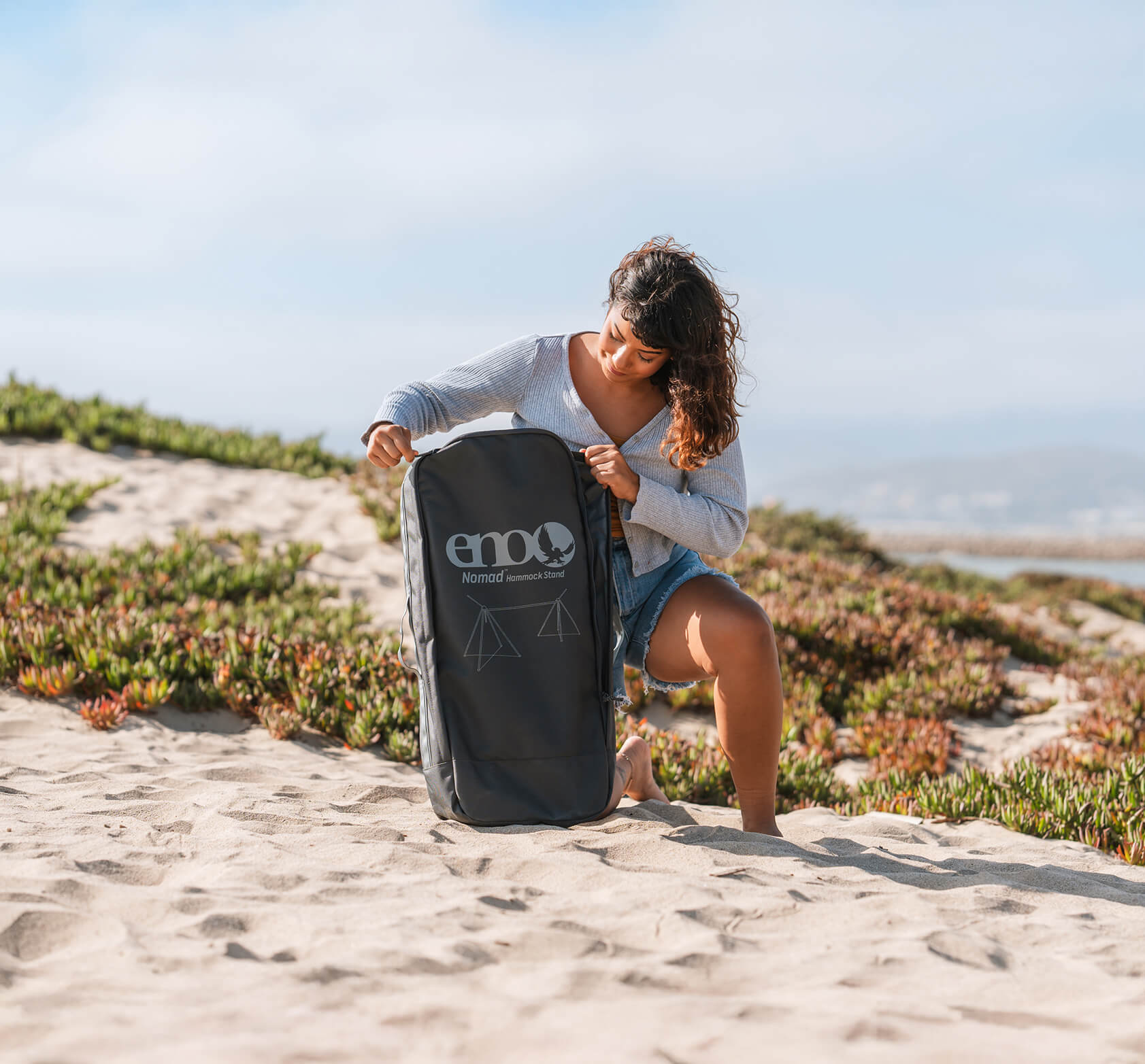 Person sitting on a sandy beach opening a packed ENO Nomad Hammock Stand custom backpack.