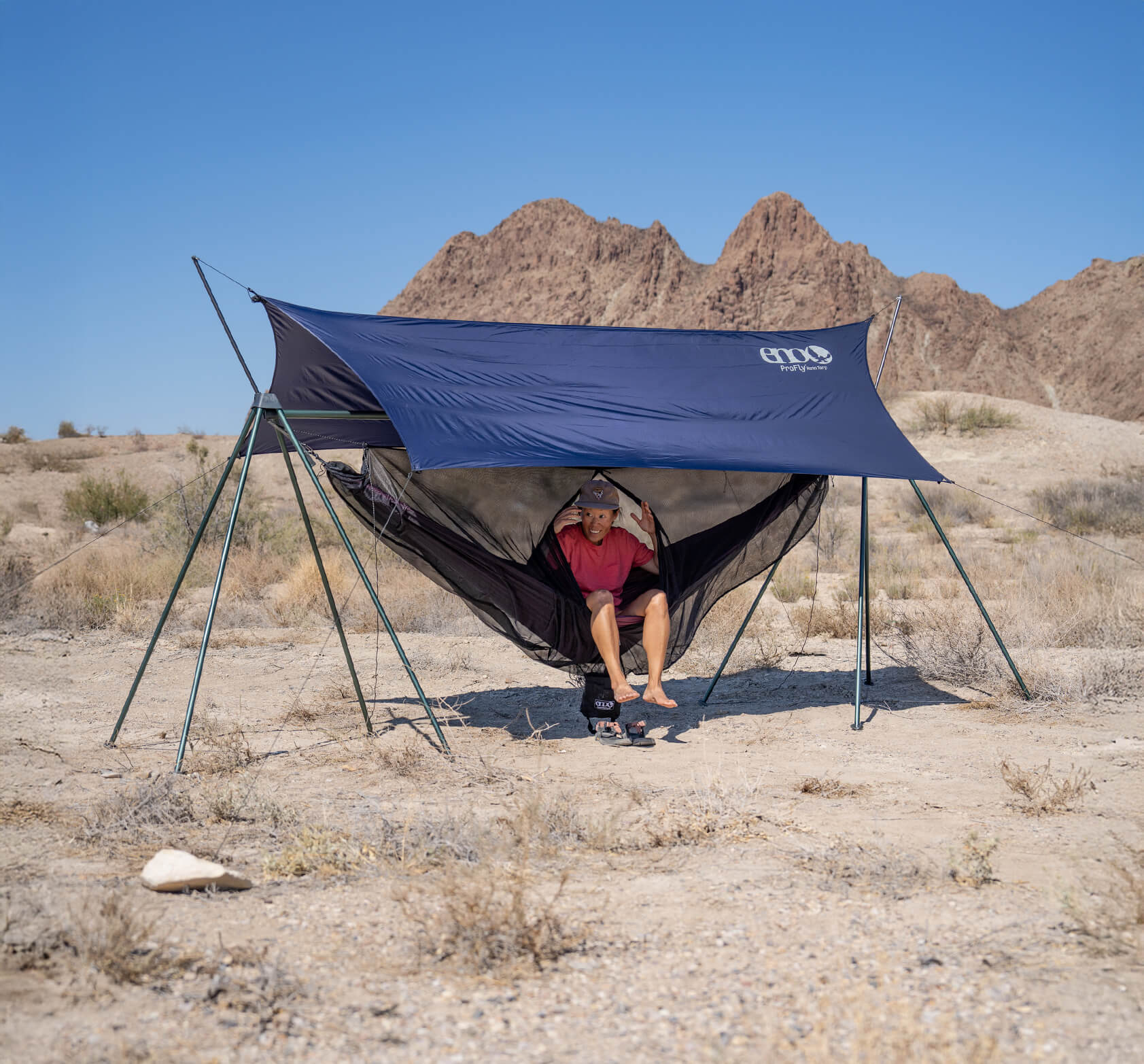 Person using the Nomad Hammock Stand with the addition of a ProFly Rain Tarp and Guardian Bug Net in a desert landscape.