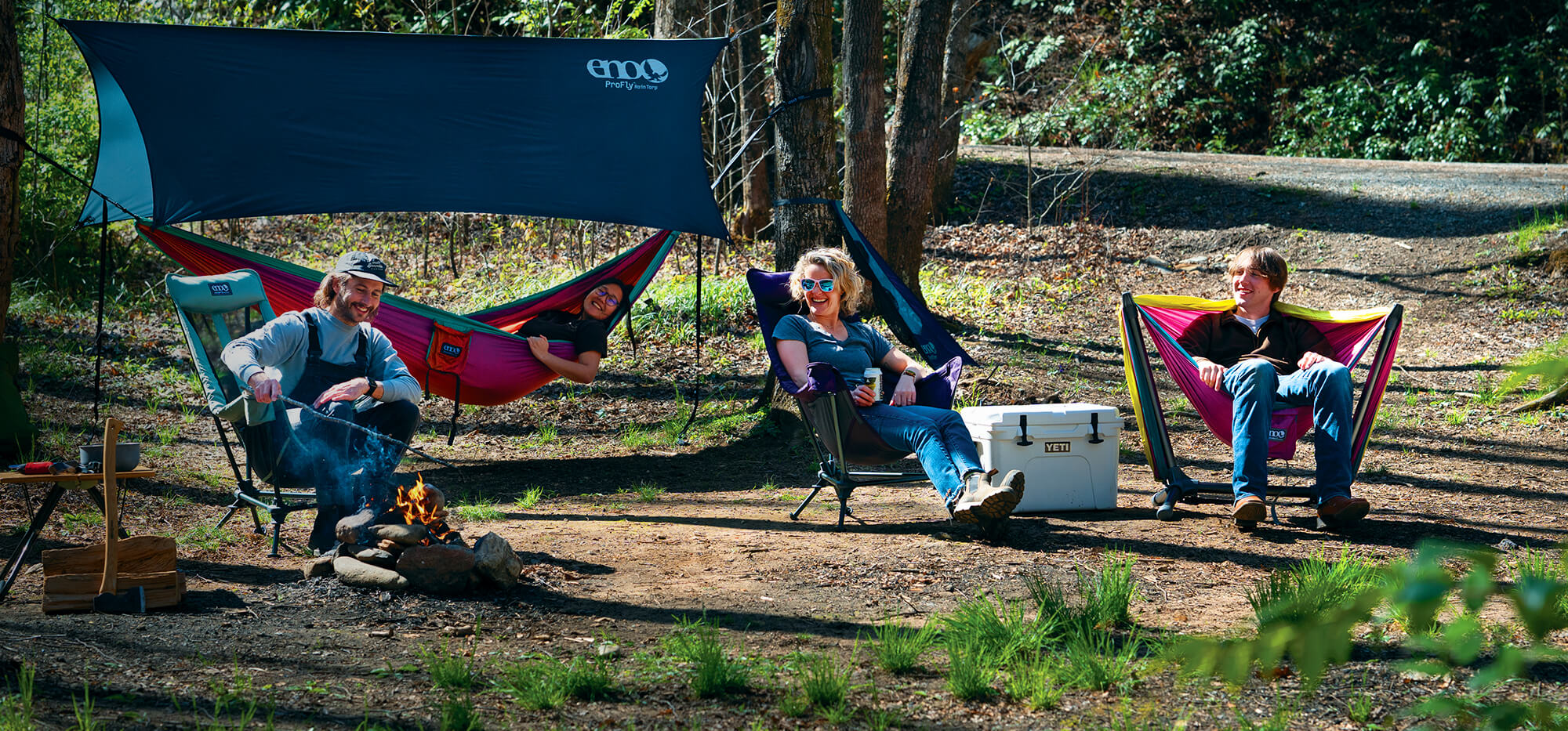 A group of friends sit fireside at their camp site laughing while relaxed in the ENO Lounger DL Mesh Chair, Lounger DL Chair, Parklite Hammock Chair Stand with DoubleNest Hammock, while a DoubleNest Hammock Print under a ProFly Rain Tarp hand g in the wooded background.