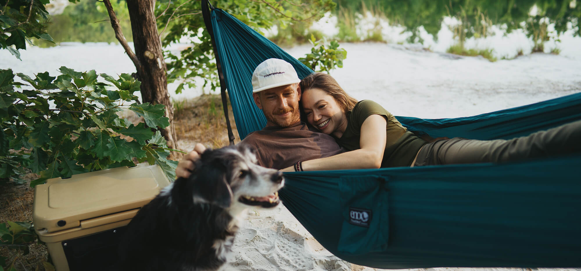 Two people laying in a ENO hammock while petting a dog in a beach setting.