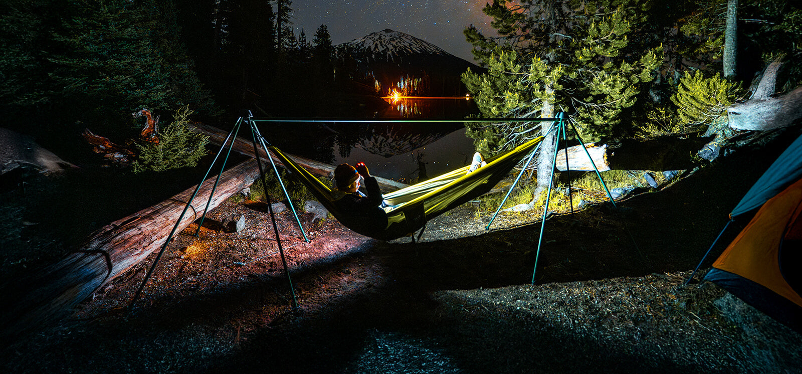 A person laying in a ENO DoubleNest Hammock set up on a ENO Nomad Hammock Stand with their headlamp on in a night time setting with mountains in the background.