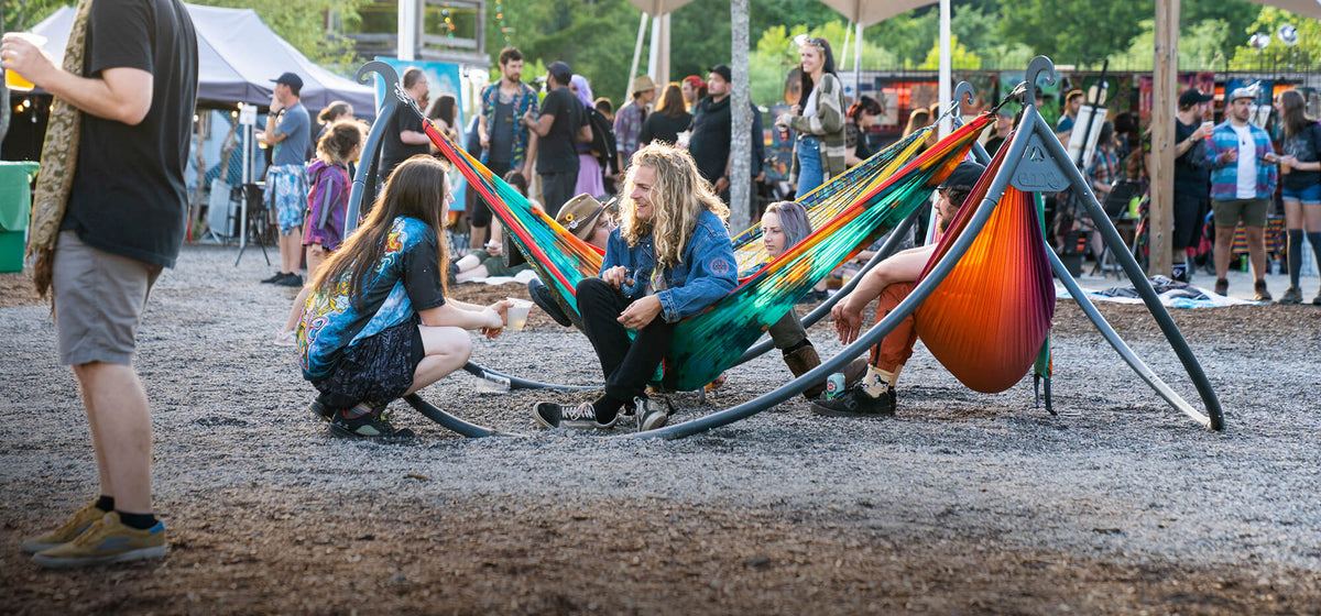 People relaxing in colorful ENO DoubleNest Print Hammocks attached to the ENO ENOpod Hammock Stand at a festival with a casual atmosphere.