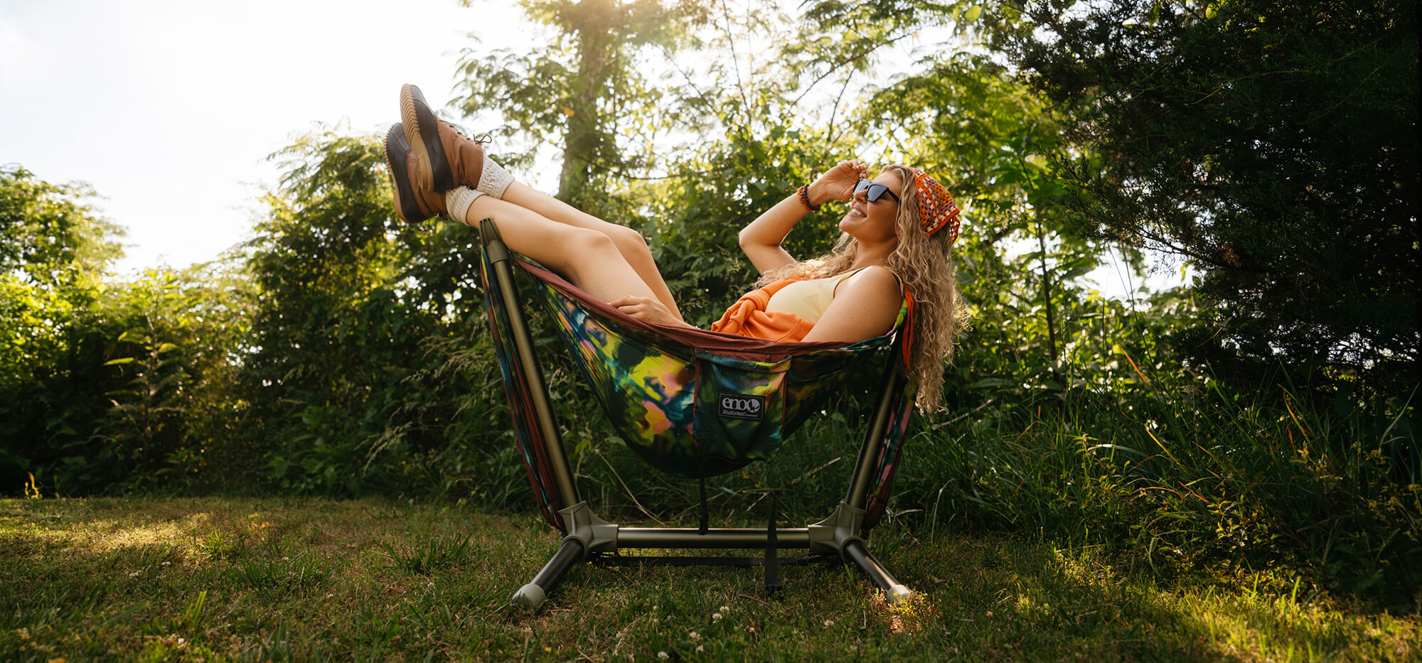 A person sitting with their legs in the air in an ENO DoubleNest Hammock Print set up on the Parklite Hammock Chair Stand.