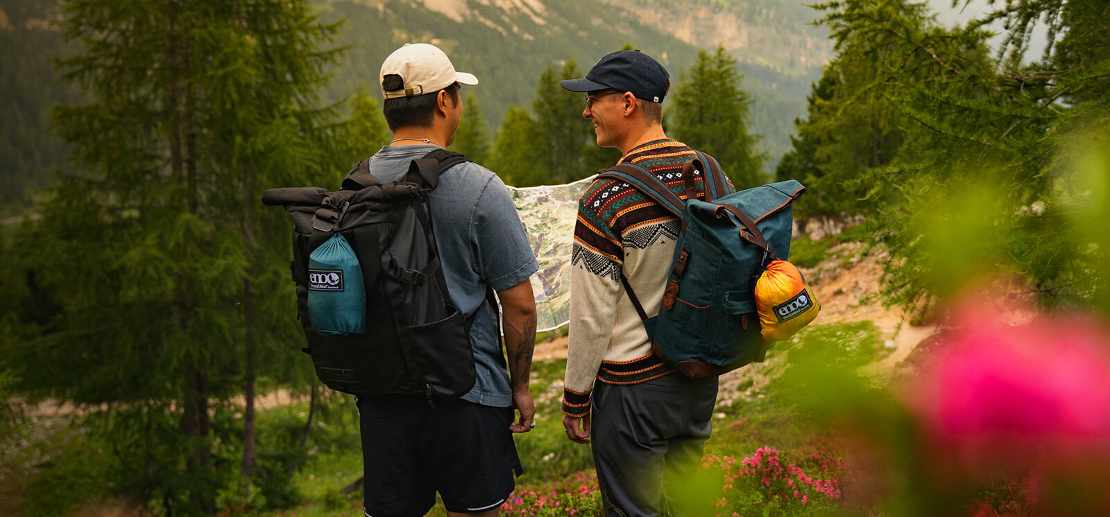 Two hikers with backpacks with packed ENO Hammocks attached, standing in a forested area with mountains in the background.