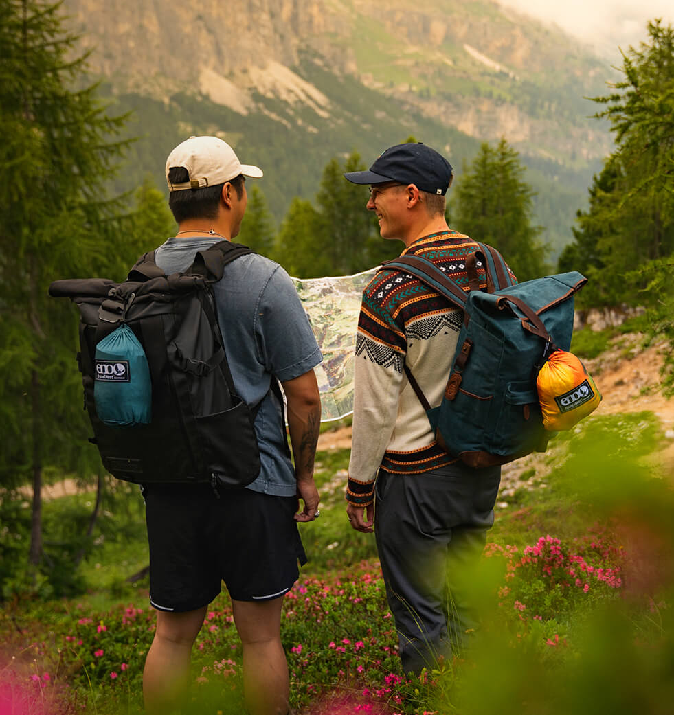 Two hikers with backpacks with packed ENO Hammocks attached, standing in a forested area with mountains in the background.