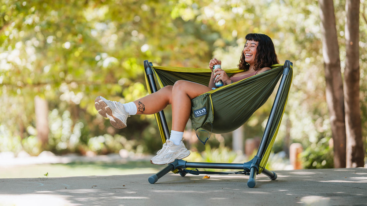 Young woman smiling kicking legs up while relaxing in an ENO Parklite Hammock Chair Stand with soft foliage in background.