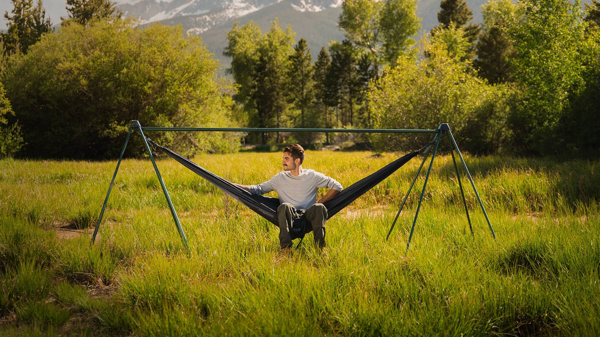 Young man sits in an ENO hammock hanging on the Nomad Hammock Stand in grassy field with trees and mountains in the background.