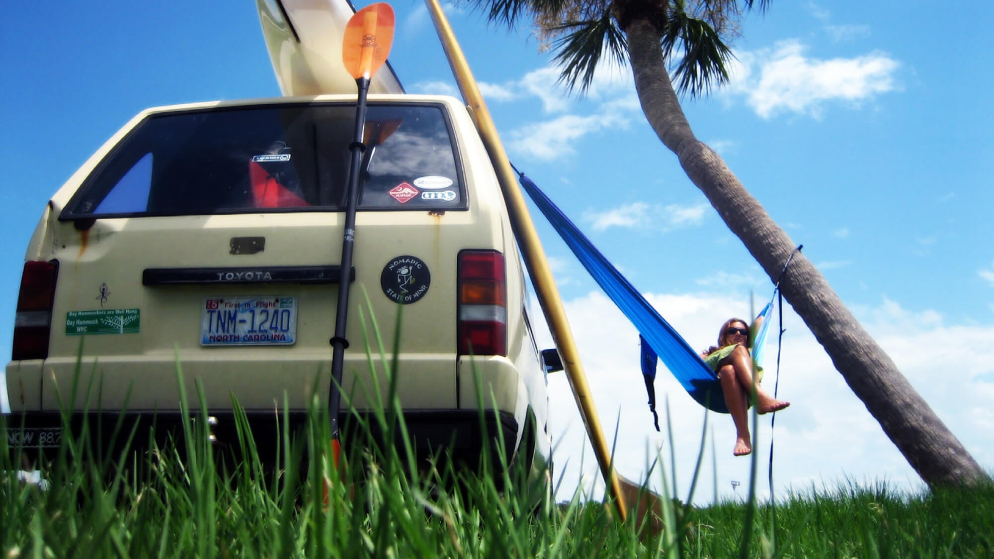 A woman sits back relaxing in an ENO portable hammock hanging between a car and tree under a blue sky.