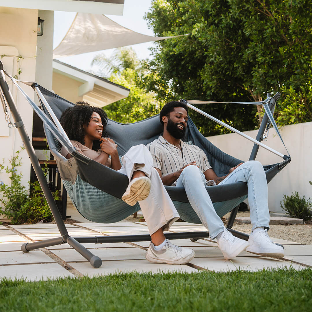 A couple sitting, laughing, in the ENO DoubleLoft Hammock Chair connected to an ENO Parkway Adjustable Hammock Stand in a backyard setting.