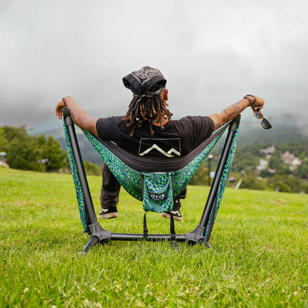 Person sitting in an ENO DoubleNest Hammock Print in Optical Illusion set up on a Parklite Hammock Chair Stand with a scenic background