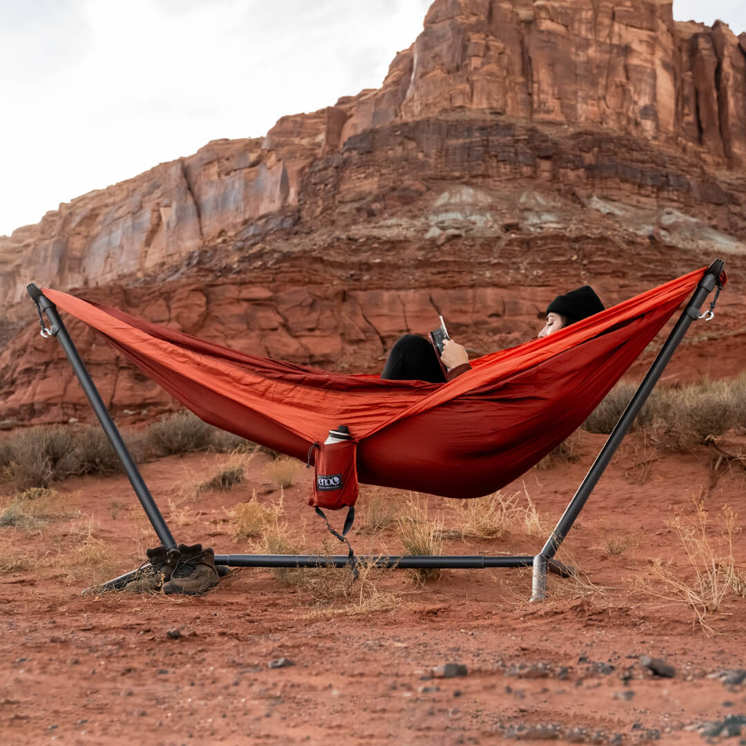Person lays back reading in an ENO DoubleNest Hammock in Canyon Coral set up on Parkway Hammock Stand in front of desert landscape.