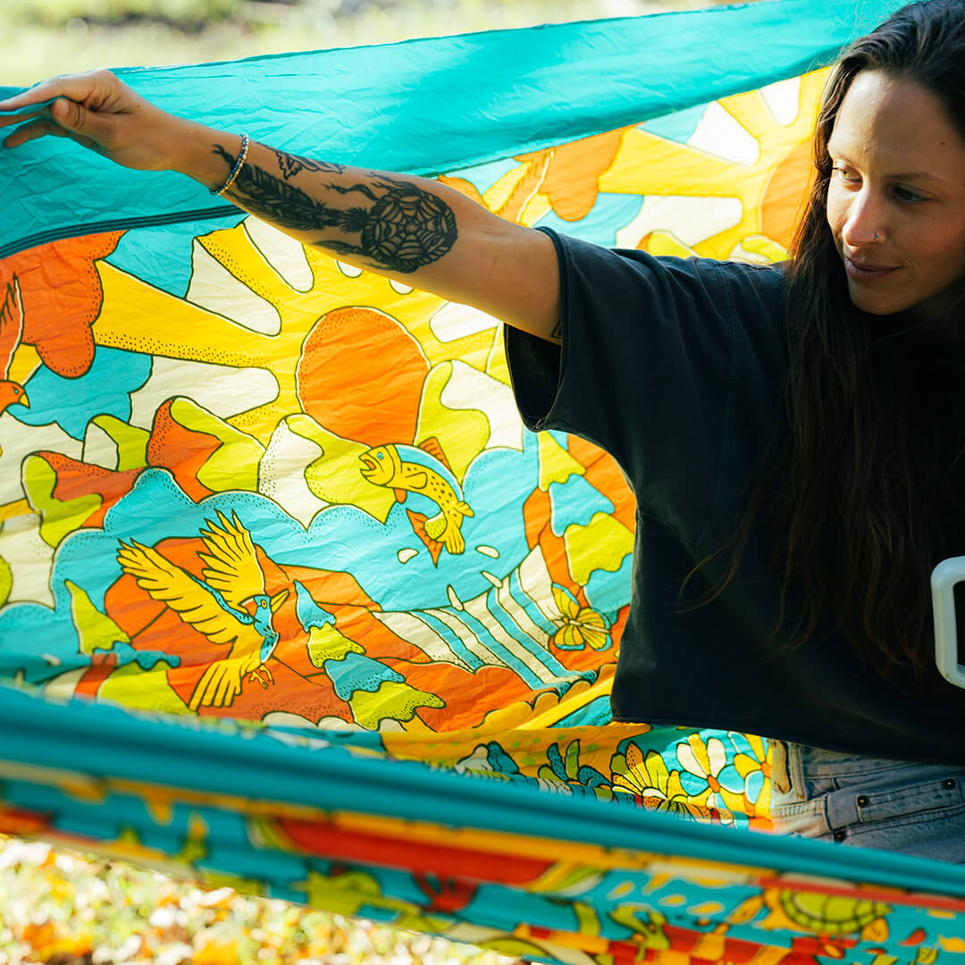 Person holding a colorful fabric of the ENO Giving Back Hammock with a vibrant design of a sun, birds, and nature.