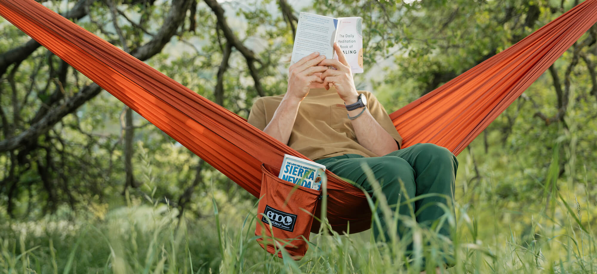 A person sits reading with a book in front of their face while sitting in an ENO TravelNest Hammock + Straps Combo surround by lush green plants.