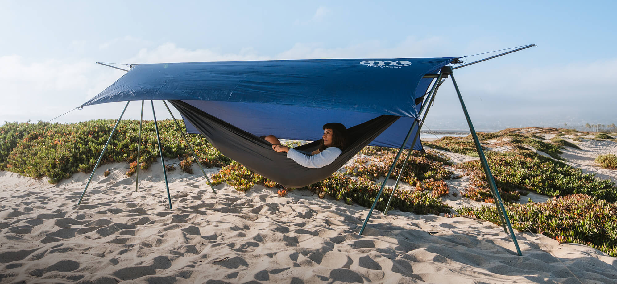 Person relaxing in a Nomad Hammock Stand under a ProFly Rain Tarp on a sandy beach.
