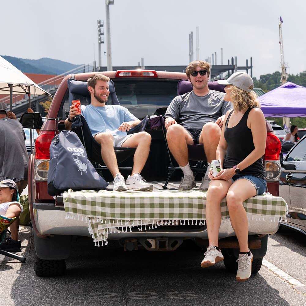 Three people sitting on a tailgate with a checkered blanket, enjoying a sunny day outdoors, while two sit in ENO Lounger DL Chairs.