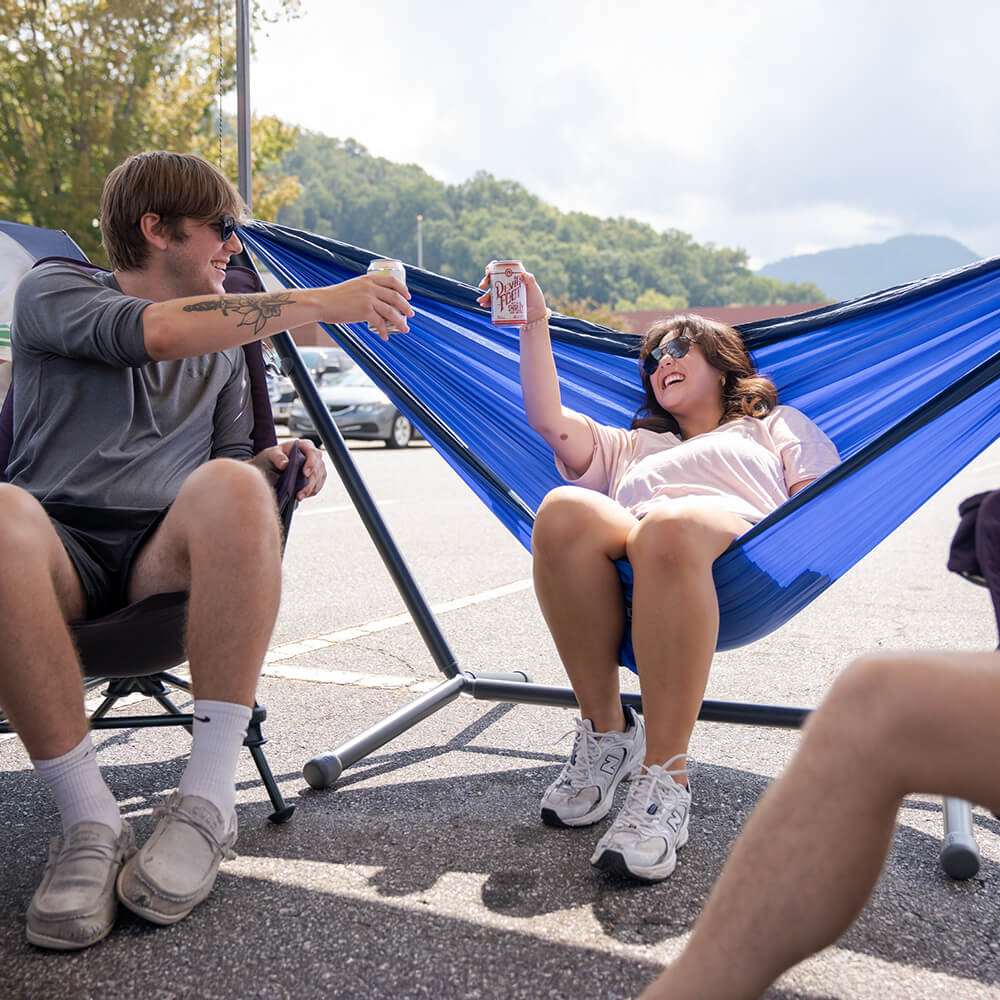 Two people sitting, cheering, in a blue ENO hammock setup on the Parkway Adjustable Hammock Stand outdoors with mountains in the background.