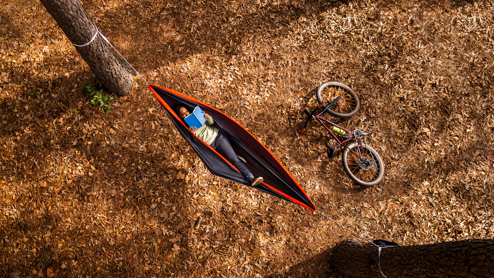 Areal view of a woman laying back reading a book in an ENO SuperSub Ultralight Hammock over leaves.