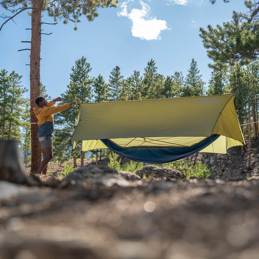 A young man sets up his ENO HouseFly Rain Tarp over their hammock setup in an open forest.