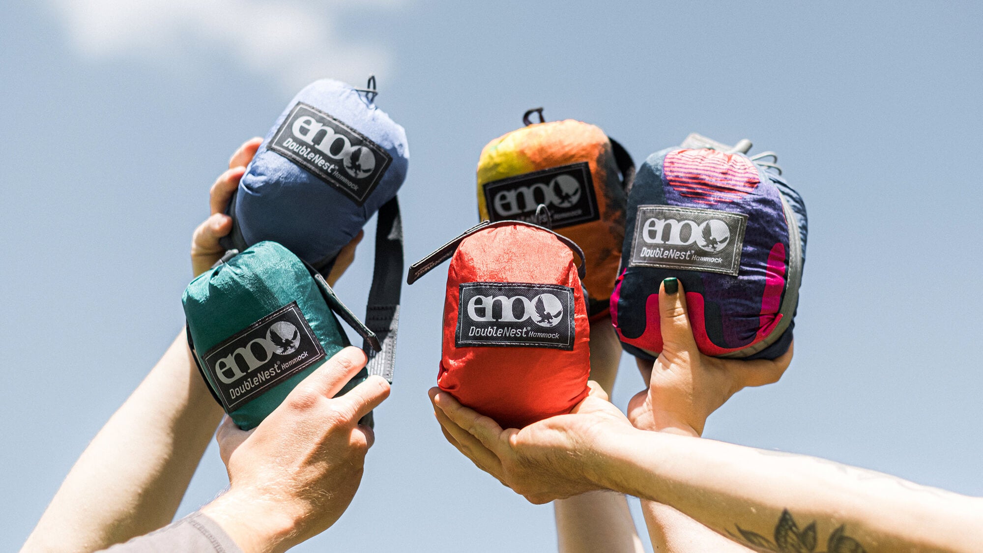 Group of arms all holding packed ENO hammock stuff sacks into the air with sky in background.