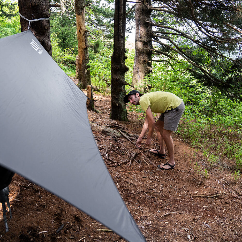 A young man stakes down an ENO ultralight hammock with a rain tarp in the forest.
