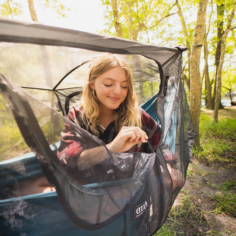 A young woman looks out into the trees in an ENO packable hammock with a hammock bug net.