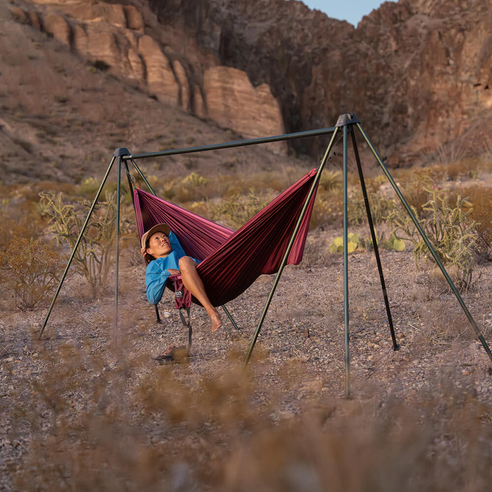 A person lying in an ENO adventure hammock set up on a portable hammock stand in a desert with mountains in the background.