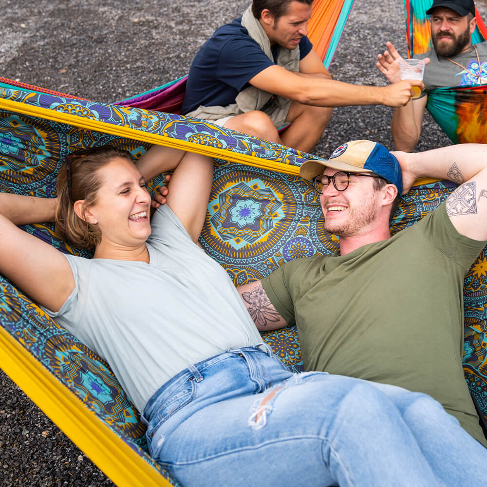 Two people relaxing in a colorful hammock with friends in the background.