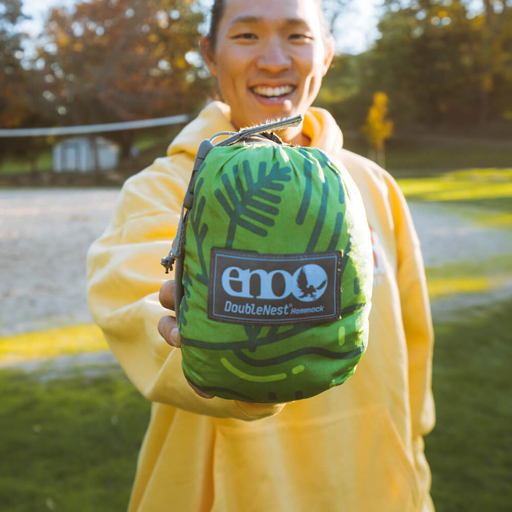 A person in a yellow sweater holding a packed ENO giving back hammock at camera.