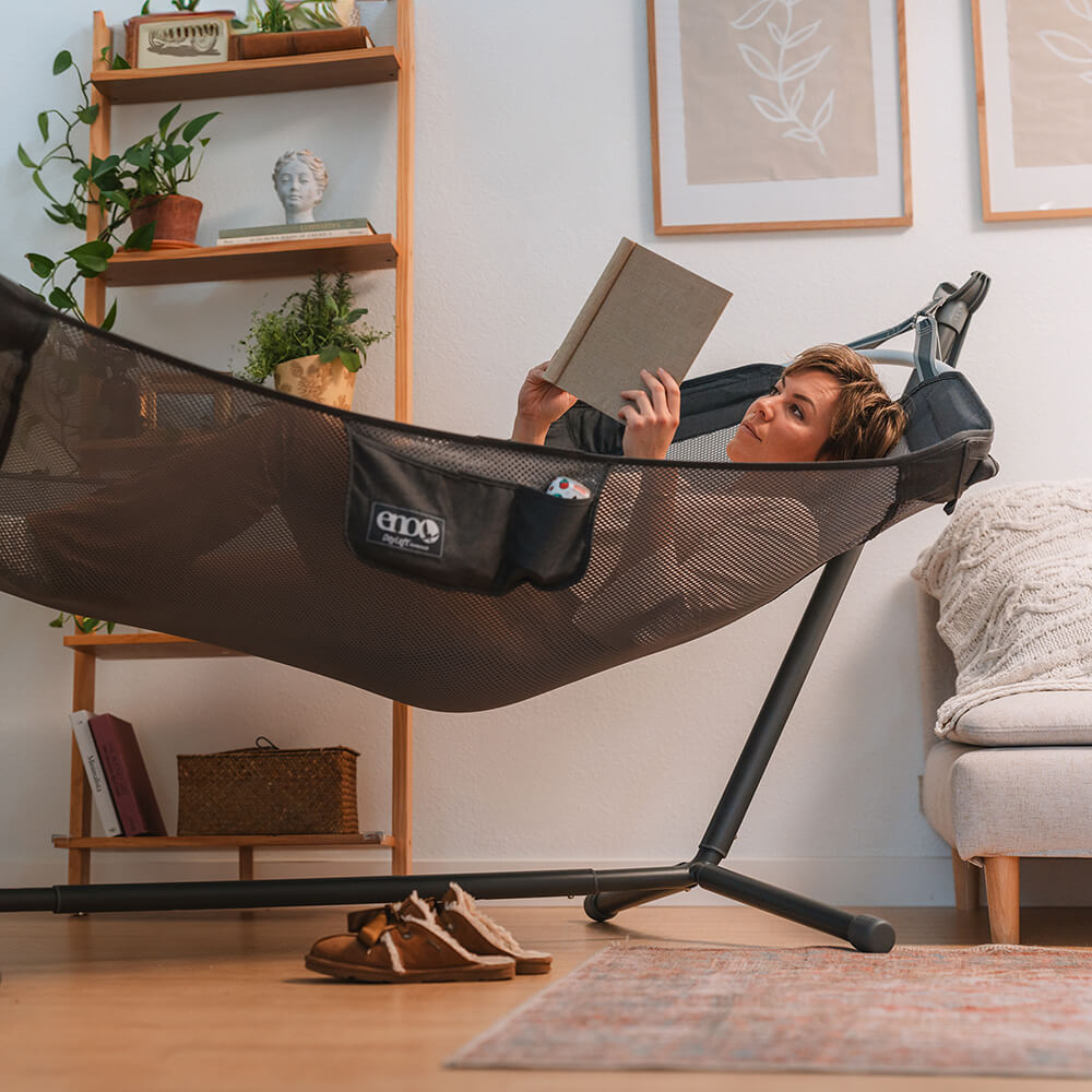 A woman lies reading in a living room with an ENO hammock stand and an indoor hammock.