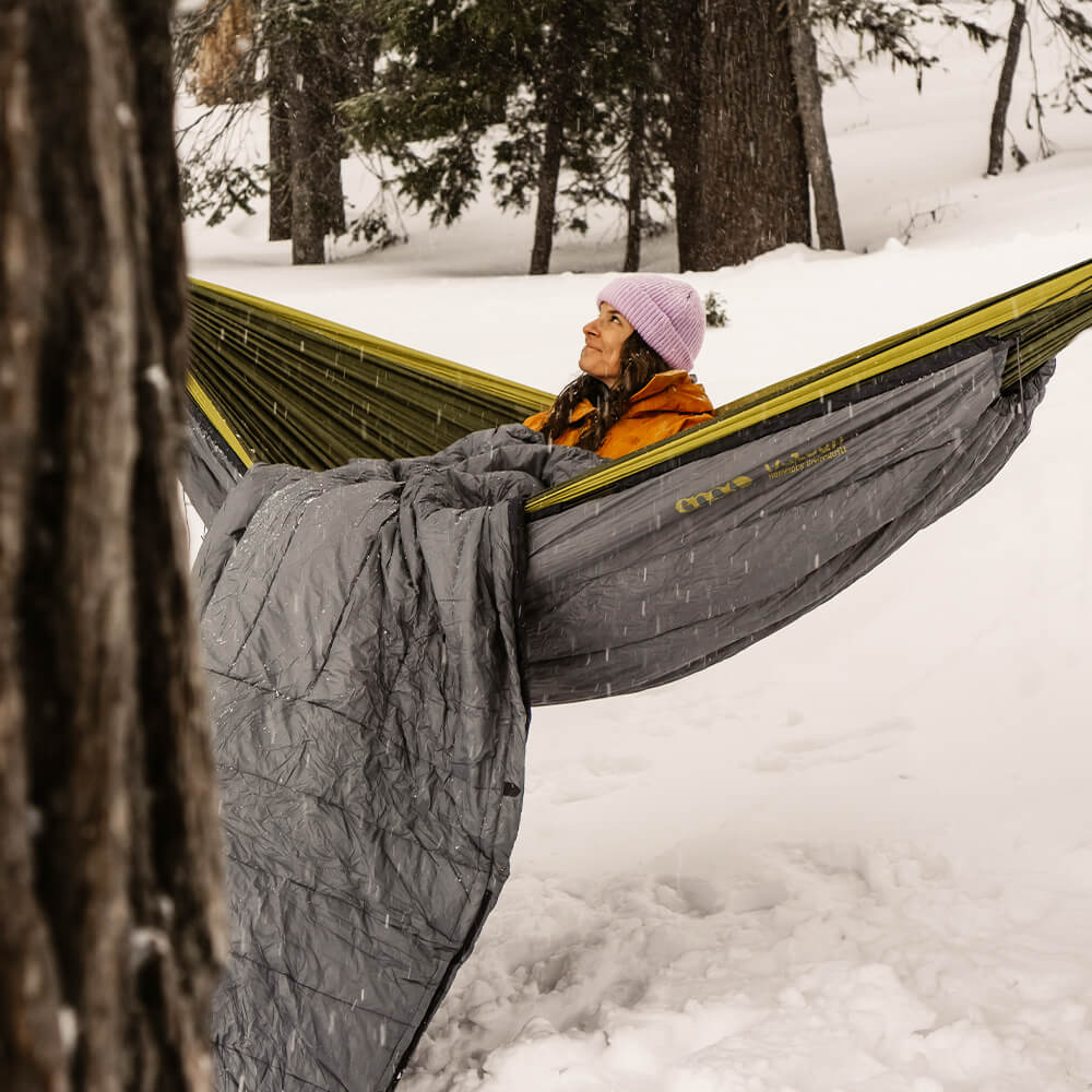 A woman looks up smiling in an ENO portable hammock and hammock insulation within the snow-covered woods.