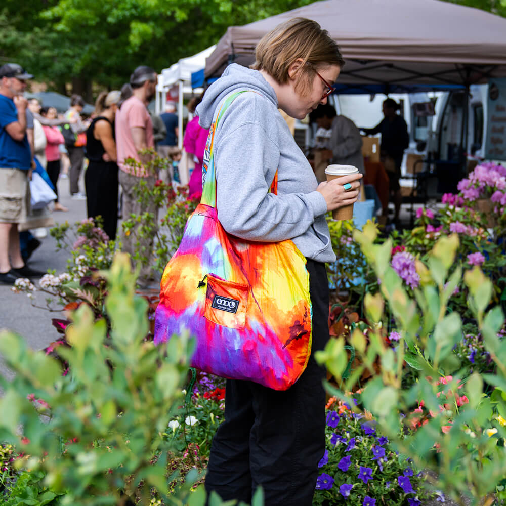 A person holding a coffee with an ENO portable tote on their shoulder while shopping at a community market.