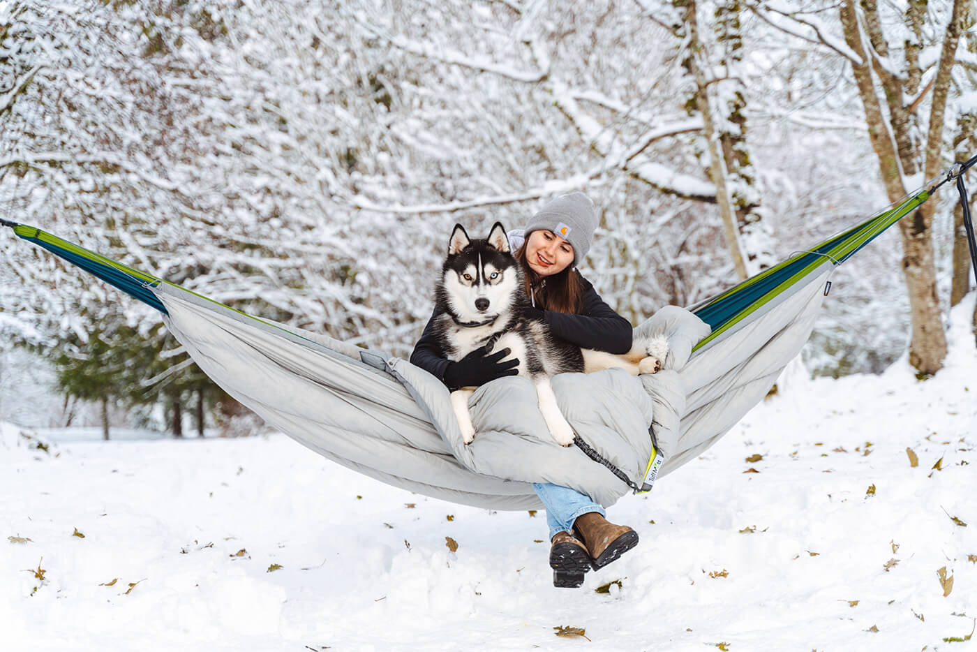 A woman and her dog stay cozy in an ENO Hammock paired with the Blaze Hammock UnderQuilt, surrounded by a snowy landscape.