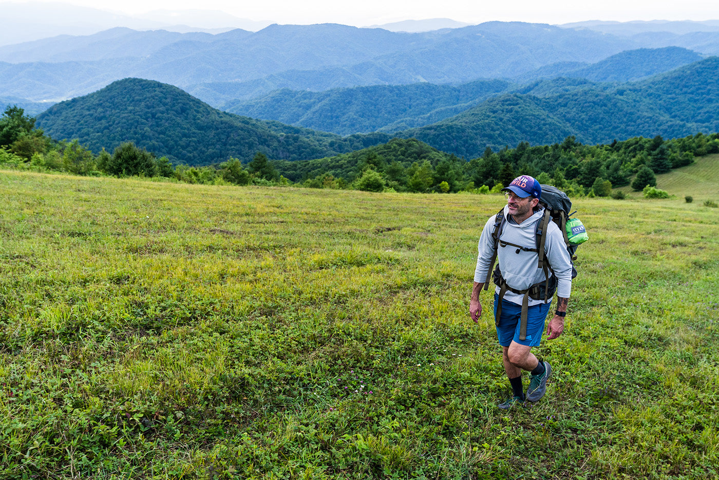 A man walking up a field with camping gear on his back and mountains in the background