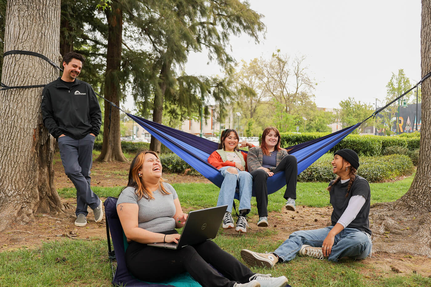 A group of friends hanging out. Two are in an ENO Hammock. One is leaning against a tree. Two are sitting on the ground 