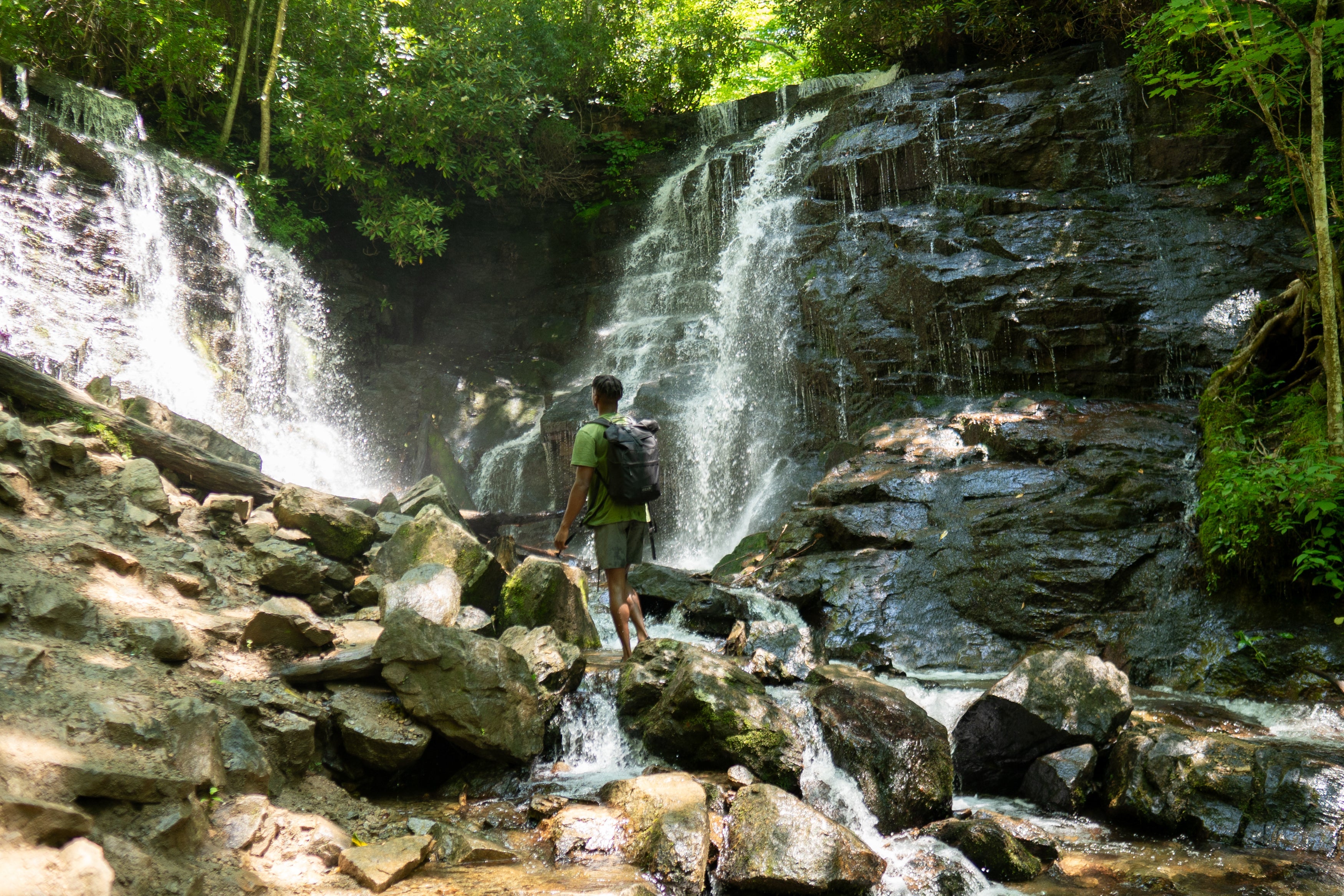 A man standing on rocks looking at a waterfall with a backpack on 