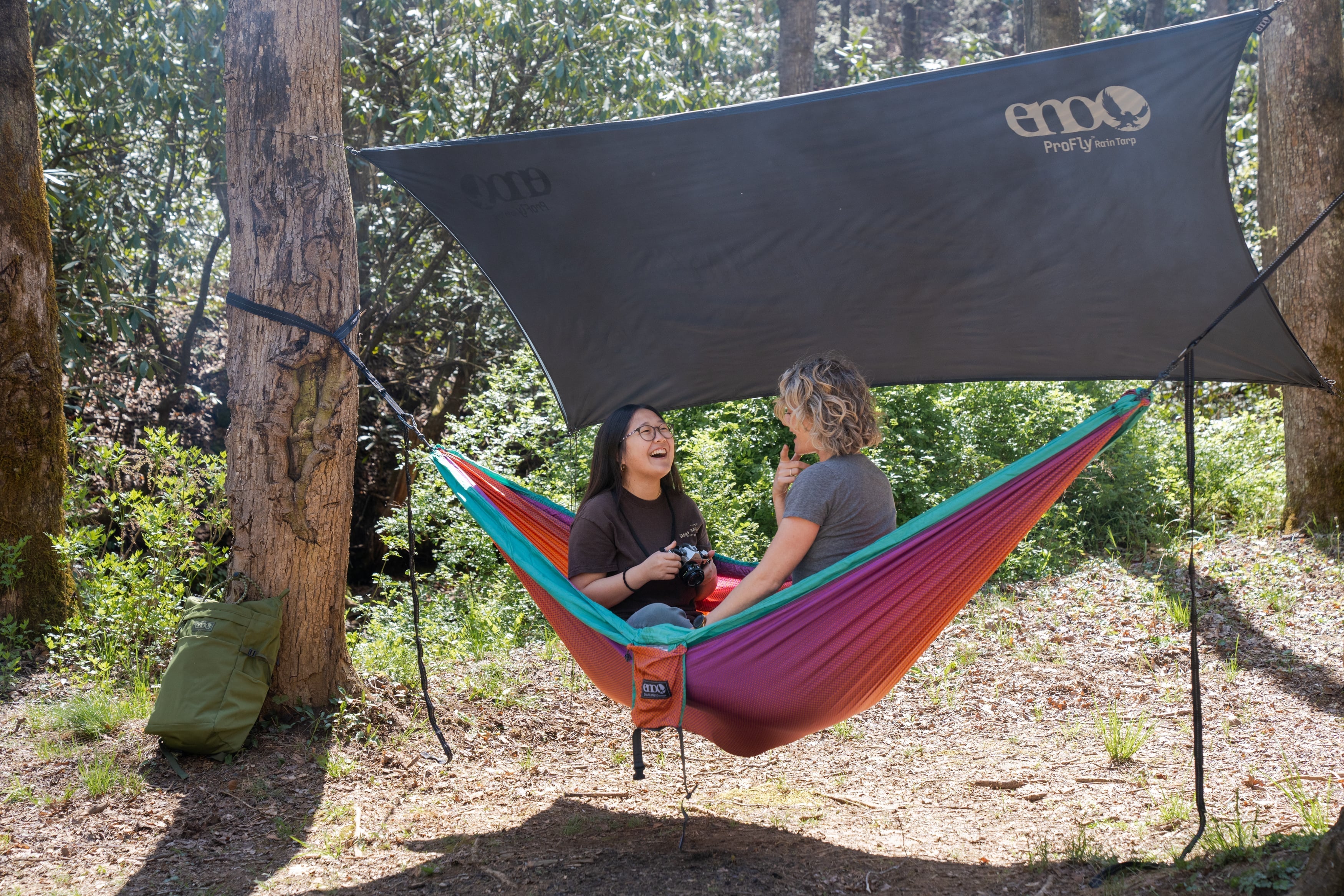 Two women in an ENO DoubleNest Hammock with an ENO ProFly Rain Tarp above them. They are at a campground and one is taking pictures of the other. 